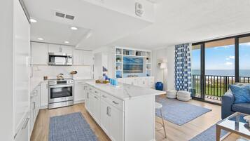 A modern kitchen with white cabinetry, stainless steel appliances, and a large island. There is a balcony with a view of the sea.