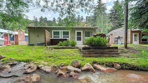 A rustic cabin with a small stream in front, surrounded by greenery and trees.