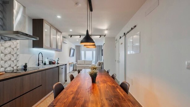 Une cuisine moderne avec une table à manger en bois, des meubles de rangement sombres et un luminaire suspendu.