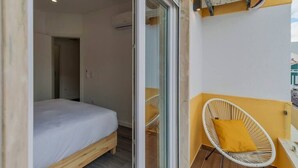 Une chambre avec un lit, une porte-fenêtre coulissante donnant sur un balcon et une chaise en osier blanche avec un coussin jaune.