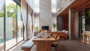 A spacious dining area with a wooden table and benches, a modern chandelier, and large windows offering views of a pool and greenery.