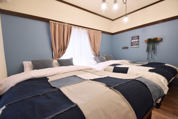 Une chambre avec deux lits, des rideaux beiges, une plante suspendue et un panneau accroché au mur.