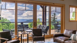 Un vaste salon doté de grandes fenêtres offrant une vue panoramique sur les montagnes, un coin salon confortable avec des canapés et des fauteuils en cuir, et une terrasse en bois à l’extérieur.