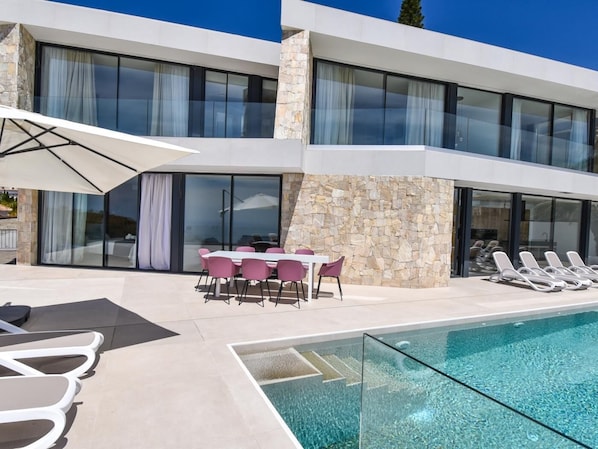 A modern house with a swimming pool, stone walls, and a dining area with pink chairs.