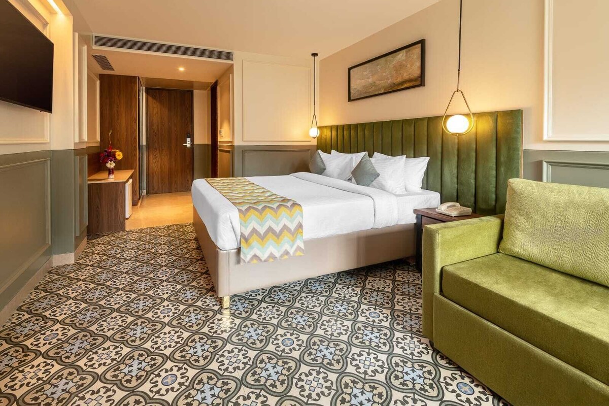 Quarto de hotel com uma cama, um sofá verde, ladrilhos estampados no chão, um quadro na parede e uma porta que dá para outro cômodo.