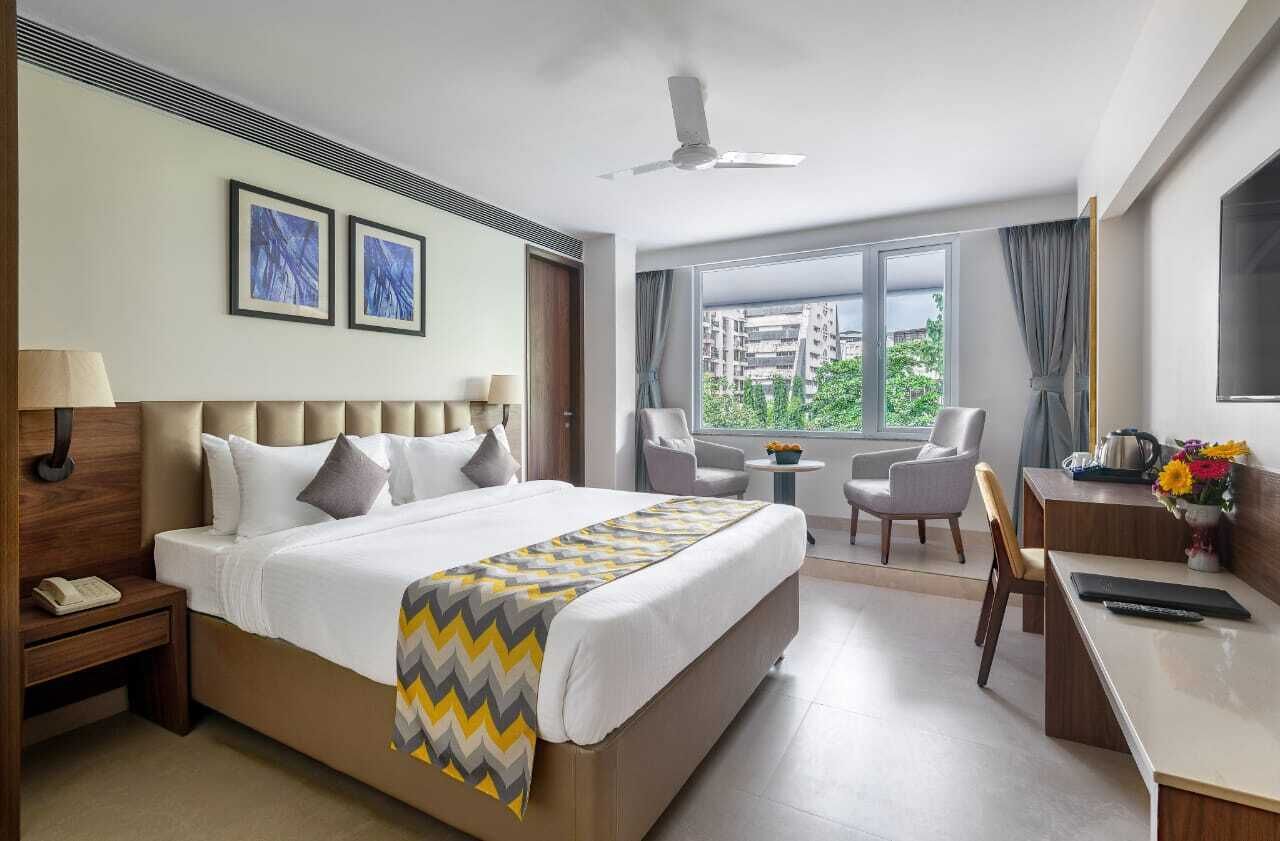 Habitación de hotel con una cama grande, un escritorio, una silla, una mesita y un ventanal con vista a la ciudad.