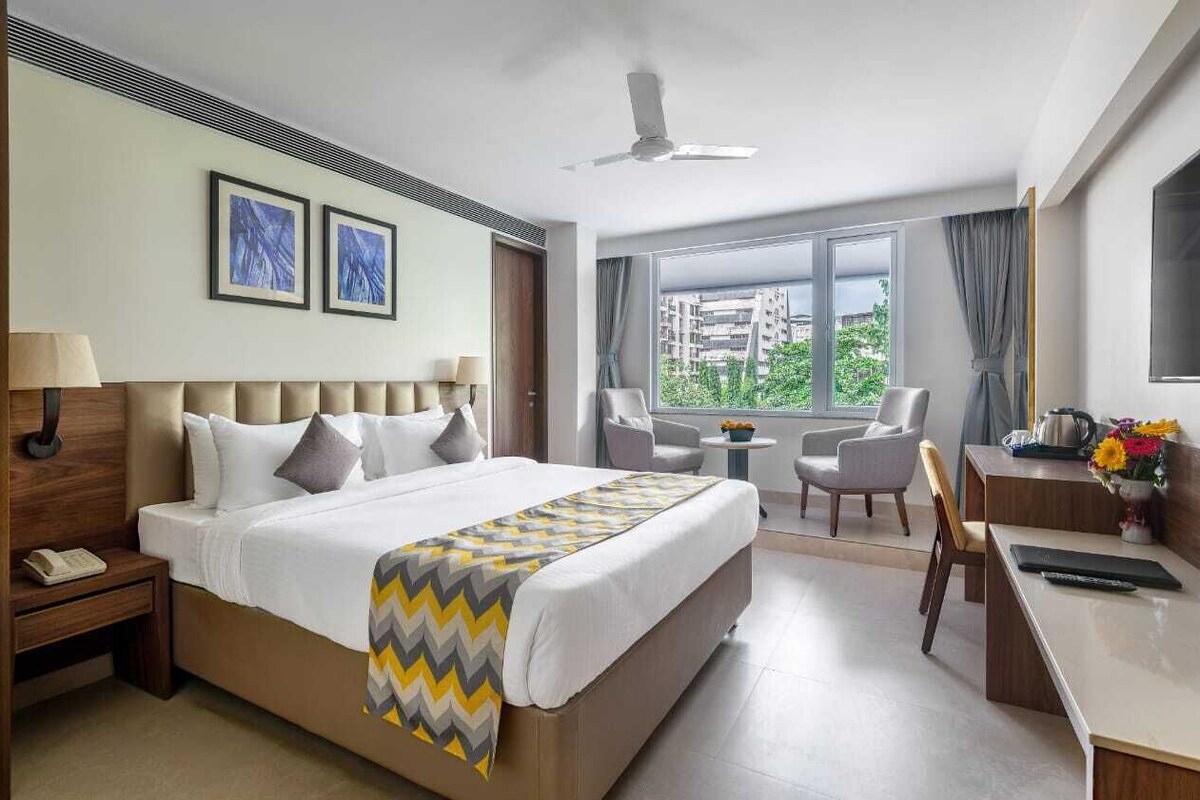 Quarto de hotel com uma cama grande, uma mesa de trabalho, uma cadeira, uma mesinha e uma janela com vista para a cidade.