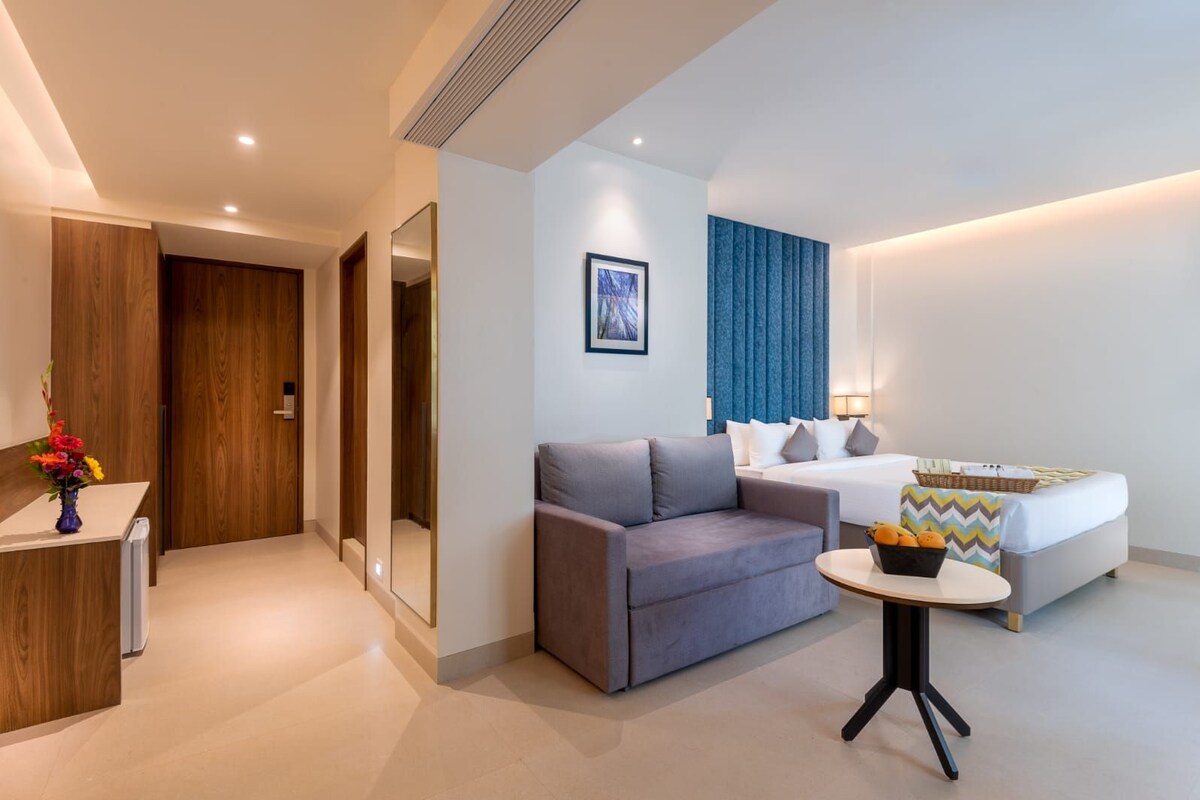 A modern hotel room with a sofa, a bed, a small table, and a desk with a vase of flowers.