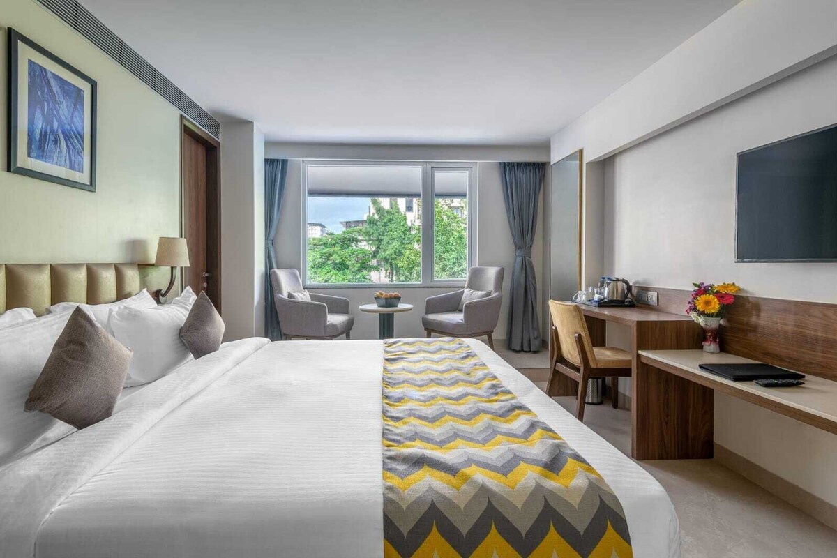 Quarto de hotel com uma cama grande, uma escrivaninha, duas cadeiras, uma mesinha, uma TV e uma janela com vista para árvores.