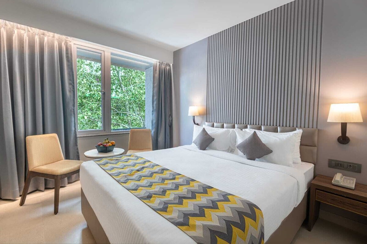 Quarto de hotel com cama, uma cadeira, uma mesinha com frutas e uma janela com cortinas.