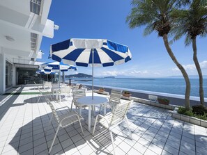 Eine Terrasse mit weißen Stühlen und blau-weißen gestreiften Sonnenschirmen, mit Blick auf ein Gewässer und Palmen.