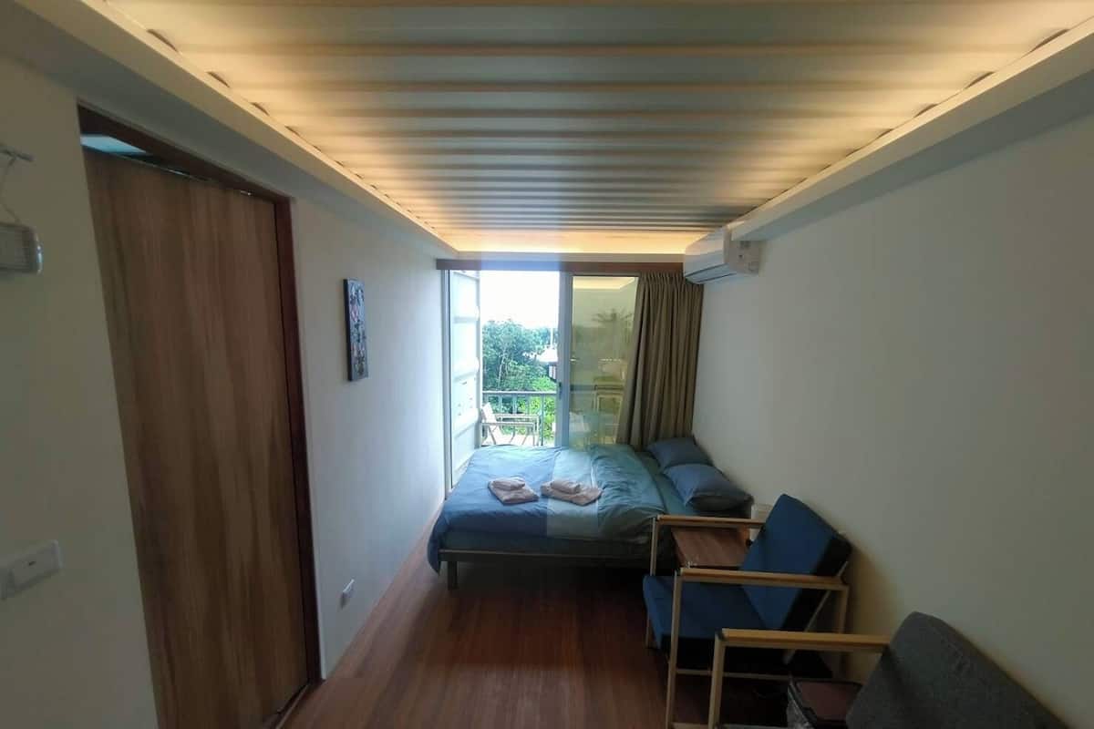 Quarto compacto com cama, uma mesinha e uma cadeira. Há uma porta de correr que dá para uma área externa com vista para vegetação.