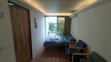 A compact room with a bed, a small table, and a chair. There is a sliding door leading to an outdoor area with a view of greenery.