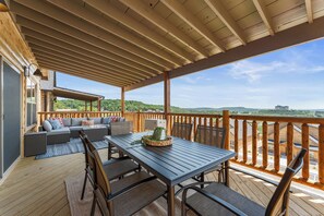A covered outdoor deck with a dining table and chairs, a sofa with cushions, and a view of a hilly landscape.