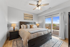 A bedroom with a large bed, two bedside tables with lamps, a ceiling fan, a view through a sliding glass door, and a wooden floor.