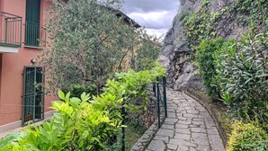 Un stretto sentiero in pietra fiancheggiato da vegetazione lussureggiante e un edificio con persiane verdi e un balcone.