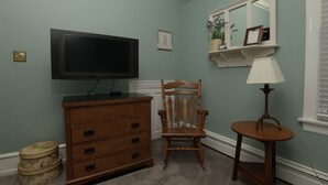 Une pièce aux murs de couleur turquoise, une commode en bois avec des tiroirs, un téléviseur posé dessus, une chaise en bois, une petite table avec une lampe et un miroir accroché au mur.