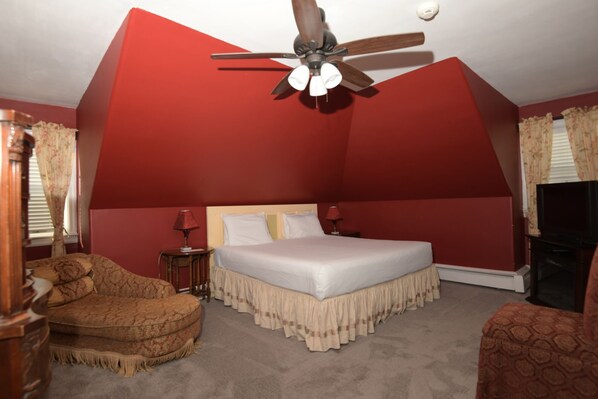 Une chambre à coucher avec un grand lit, un ventilateur de plafond, une chaise en bois et un téléviseur.