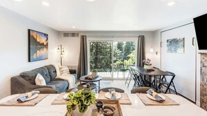 A modern living room with a sofa, dining table, and chairs, a large window with a view of greenery, and a flat-screen TV mounted on the wall. - Gorgeous Condo With River View - Pool and Hot Tub (Bend)