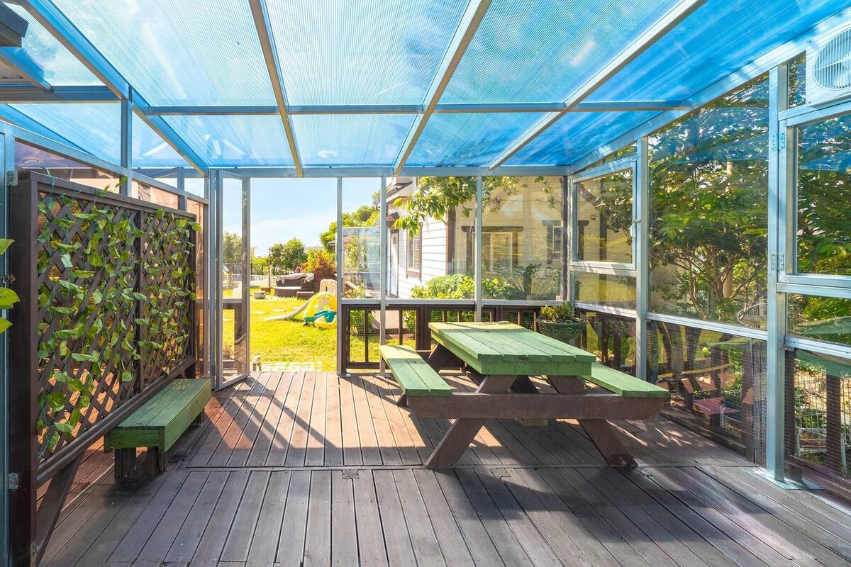 Een overdekt terras met houten vloer, groene banken en uitzicht op een speeltuin.