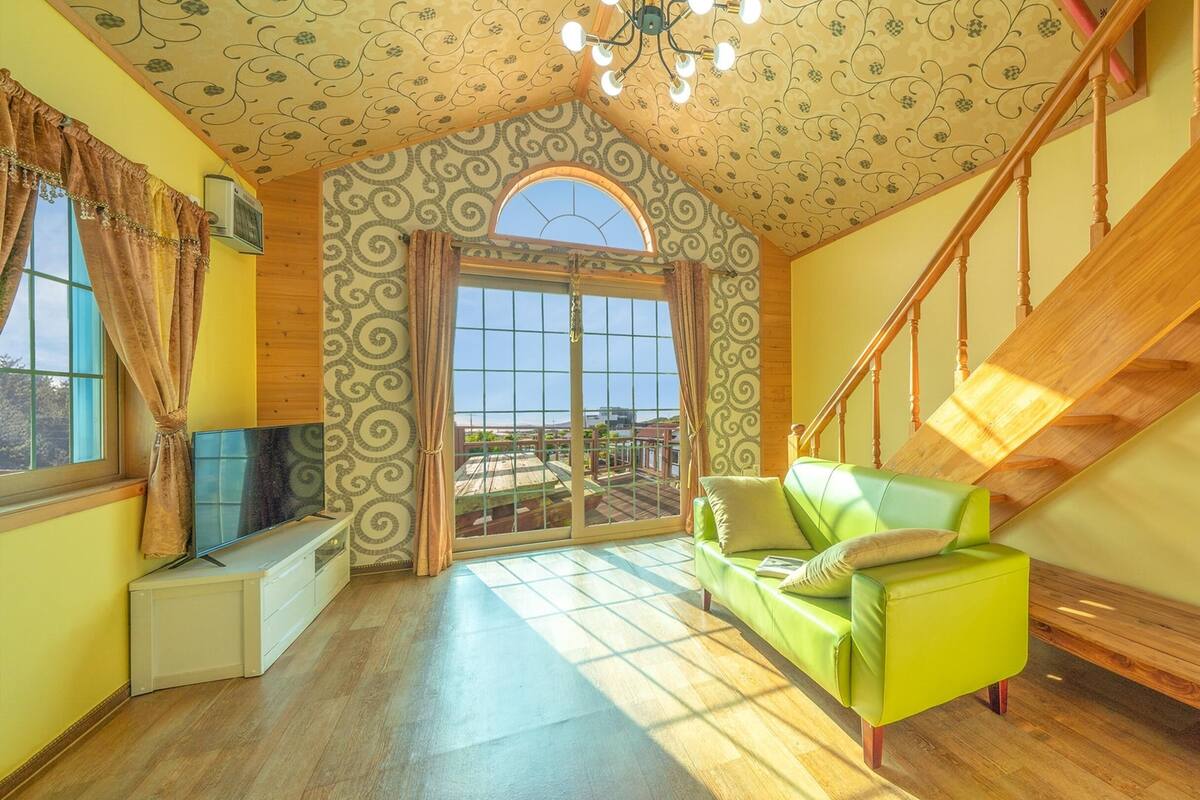 Ruime kamer met een groene bank, houten trap en een groot raam met uitzicht op de omgeving.