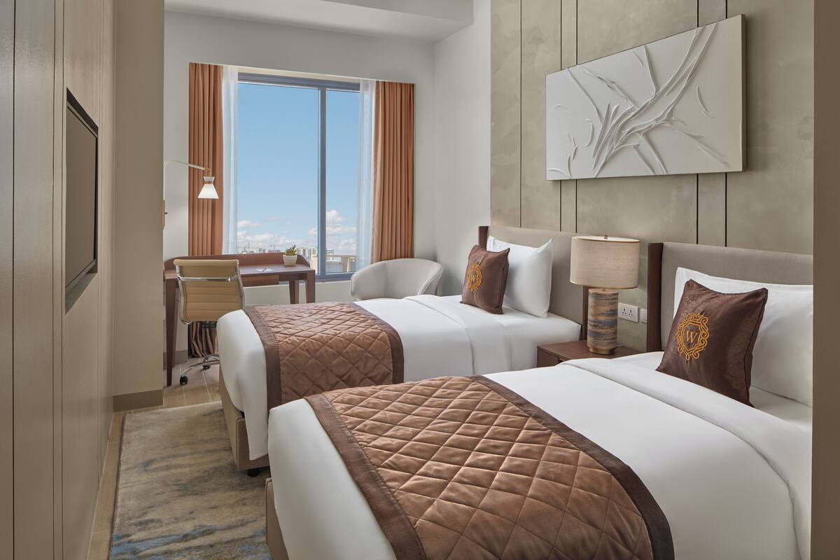 A hotel room with two beds, a desk, and a large window offering a city view.