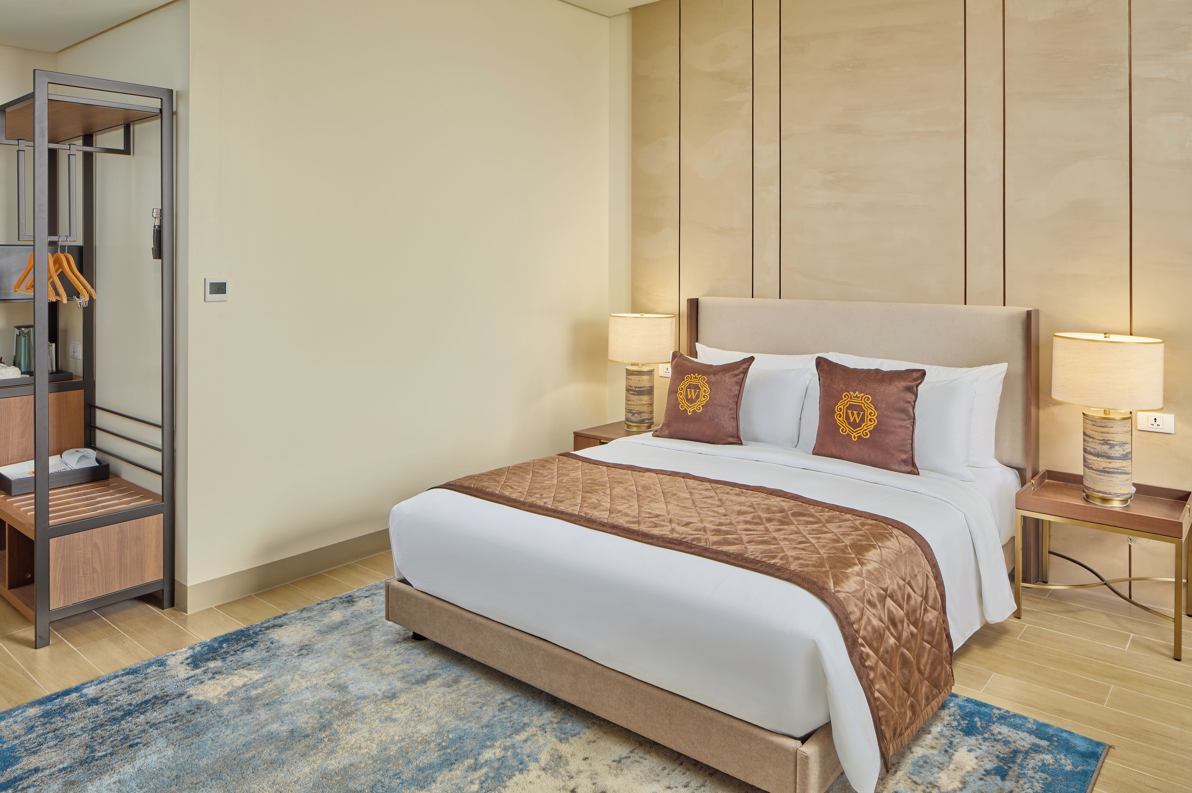 A neatly arranged hotel room with a bed, bedside lamps, a nightstand, a wooden wardrobe, and a blue patterned rug.