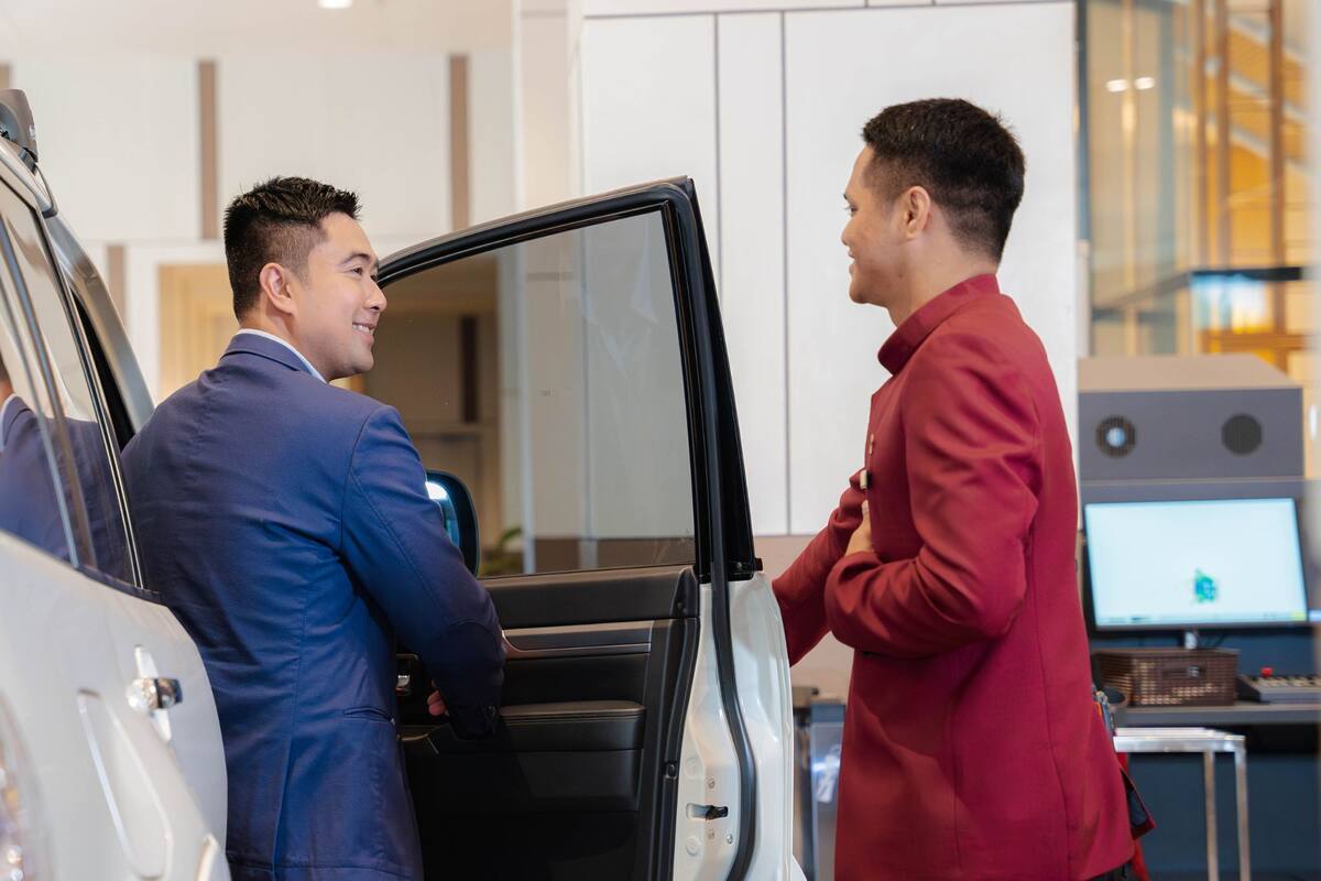 Two individuals interacting near a vehicle with an open door, in a modern indoor setting with a computer monitor in the background.
