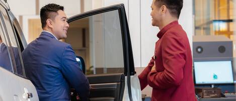 Two individuals interacting near a vehicle with an open door, in a modern indoor setting with a computer monitor in the background.
