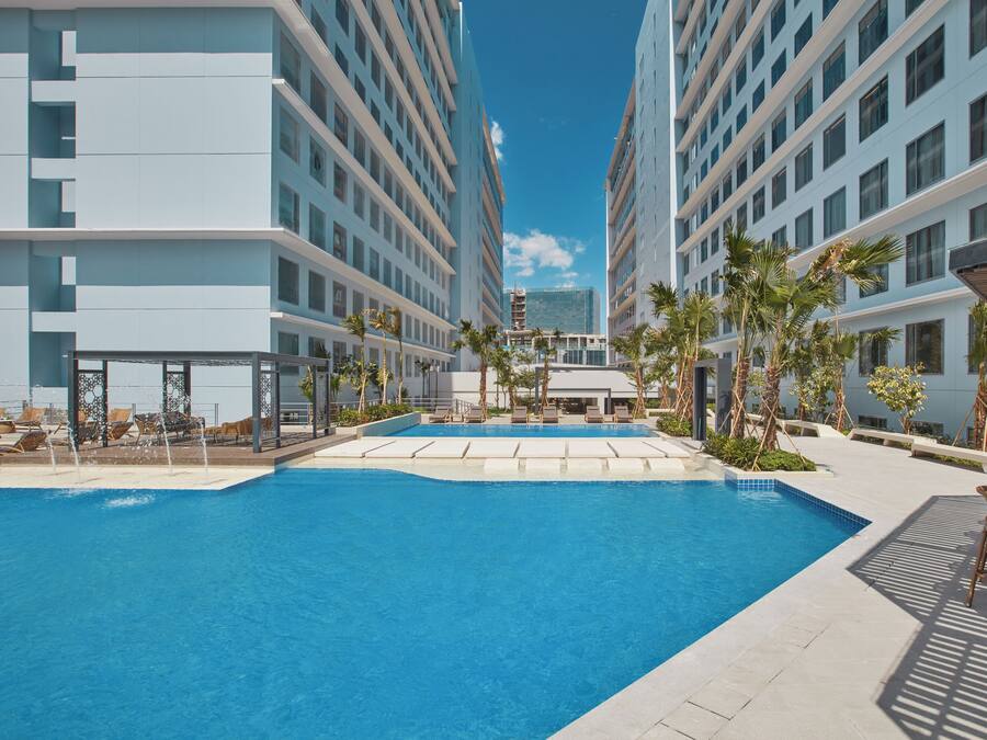 A swimming pool surrounded by modern buildings, with lounge chairs and palm trees.