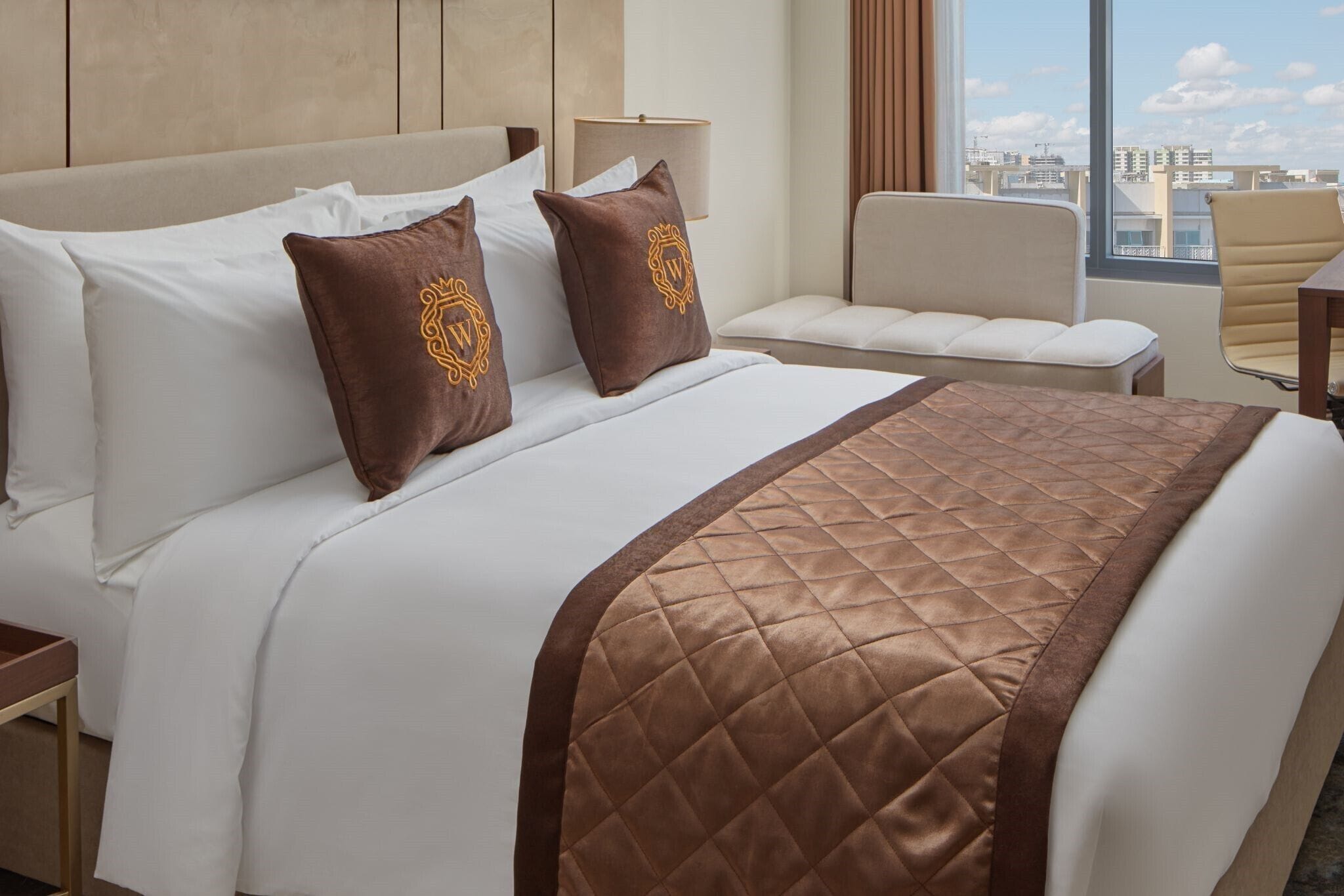 Une chambre d’hôtel avec un grand lit, deux oreillers ornés d’un logo, un couvre-lit marron matelassé, une lampe de chevet, une chaise et une vue sur le paysage urbain par une grande fenêtre.