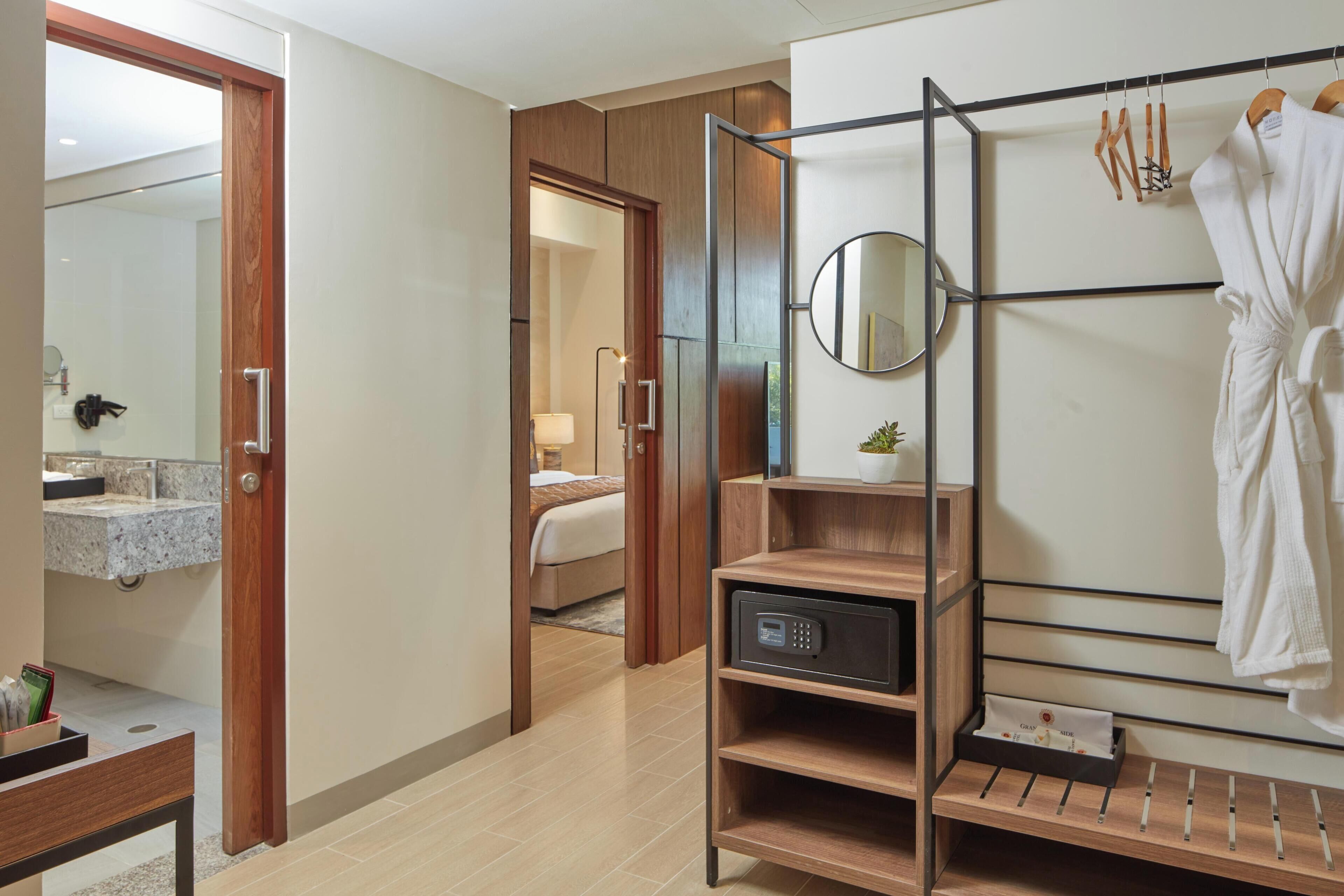 A hotel room with a wooden door, a bed, a bathroom with a mirror, and a robe rack with a robe.
