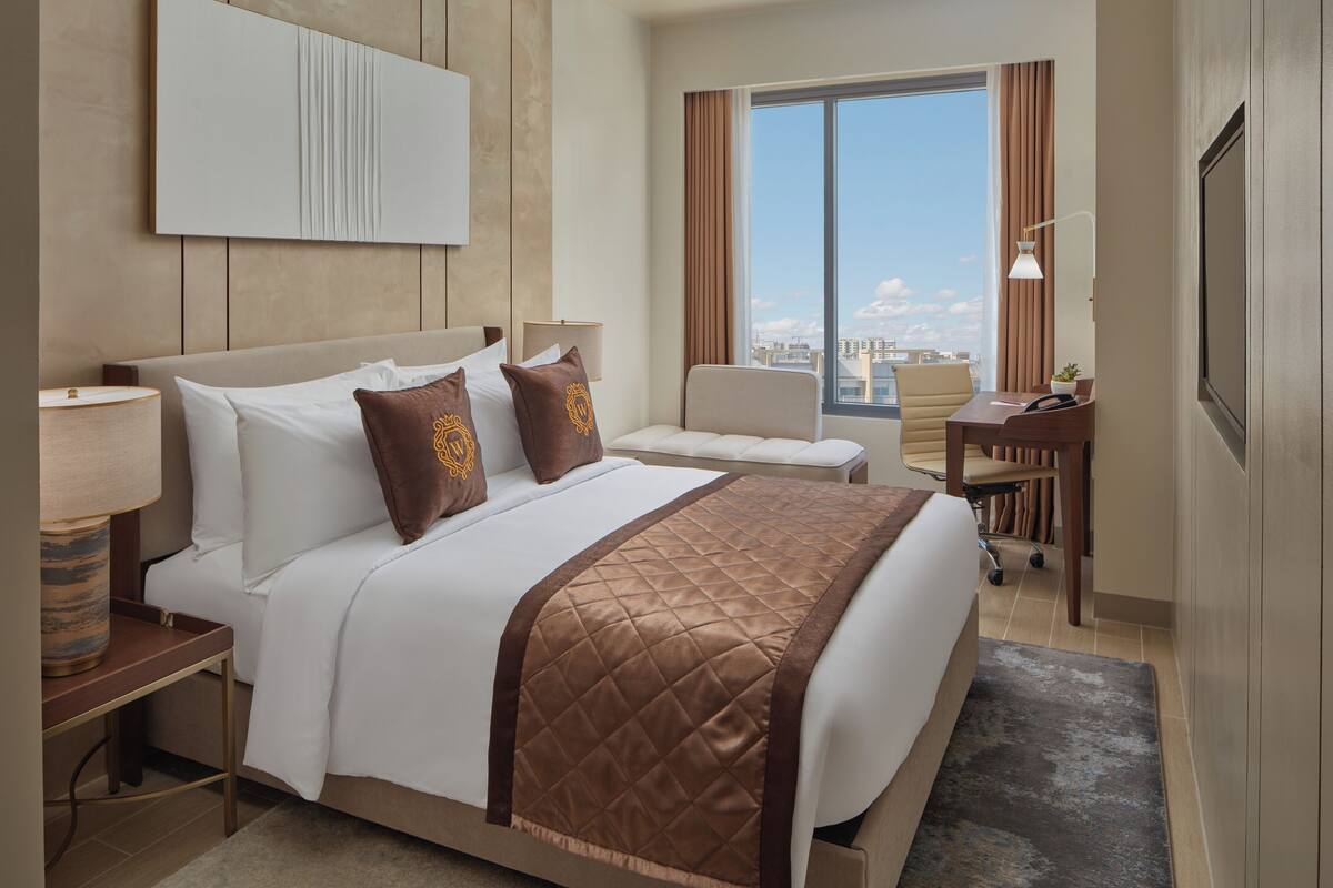 A hotel room with a large bed, a desk, a chair, and a view of the city through the window.