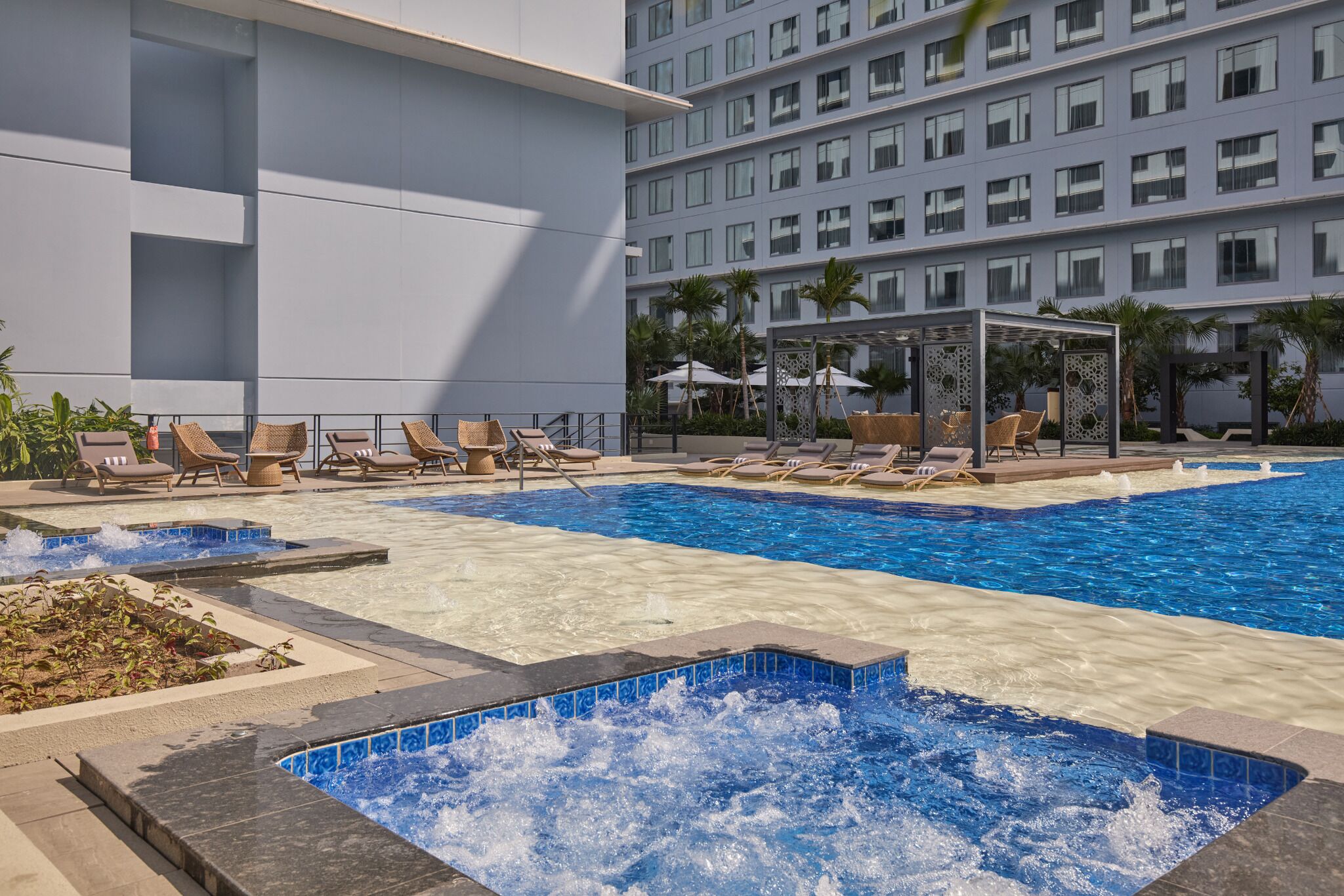A hotel pool area with multiple lounge chairs, a hot tub, and a large building in the background.