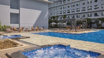 A hotel pool area with multiple lounge chairs, a hot tub, and a large building in the background.