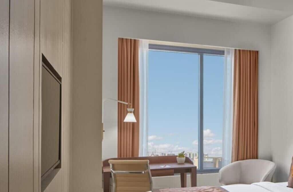 A hotel room with a large window, a desk, a chair, and a bed.
