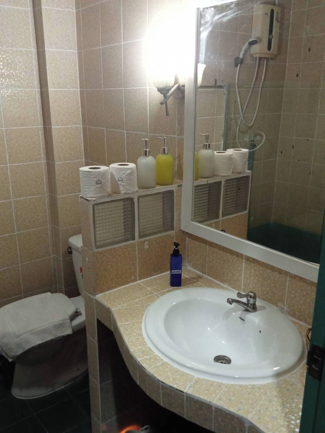 Un baño con inodoro, lavamanos y espejo. Hay artículos de aseo en la encimera y una ducha sobre el espejo.