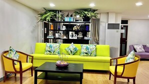 Un salón luminoso con un sofá verde, sillones amarillos, una mesa de centro negra y una estantería llena de diversos objetos.
