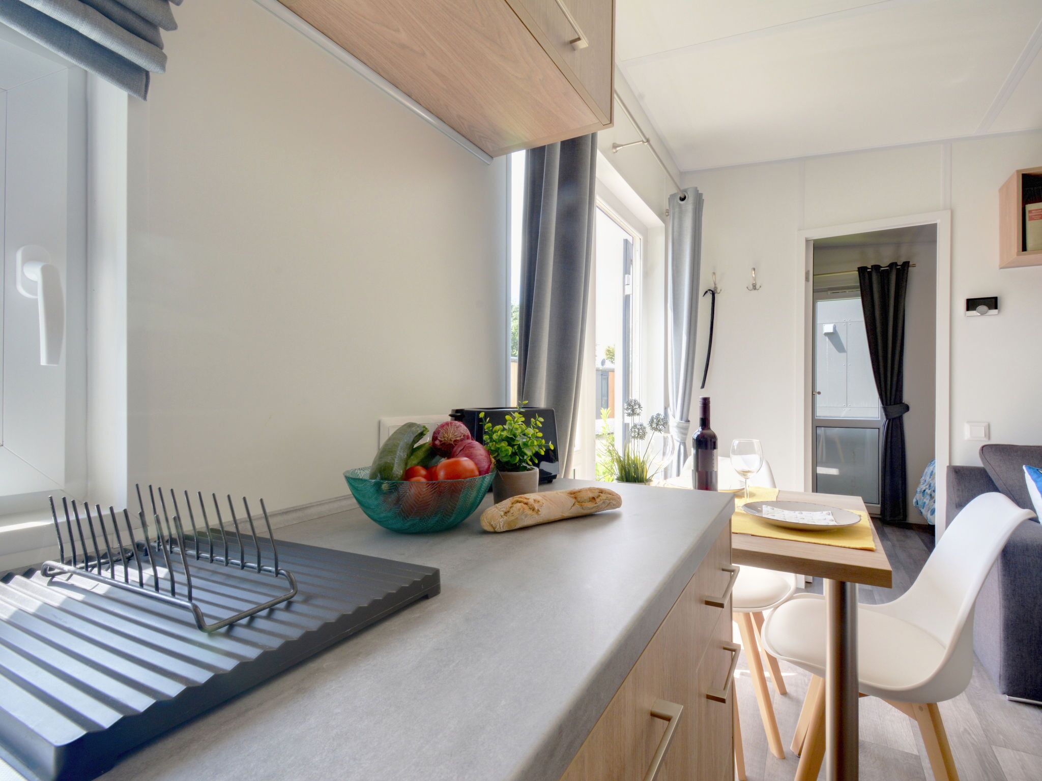A modern kitchen with a built-in oven, a fruit bowl, a wine bottle, and a dining area with a table and chairs.
