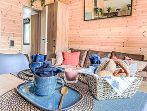 A cozy room with a wooden wall, a sofa with cushions, a table with a blue mug, a glass of drink, and a basket of bread.