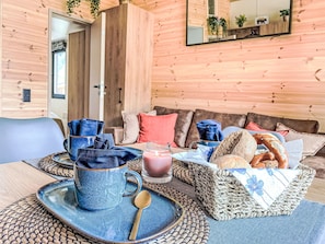 A cozy room with a wooden wall, a sofa with cushions, a table with a blue mug, a glass of drink, and a basket of bread.