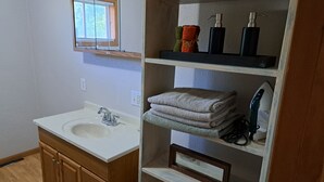 A bathroom with a sink, wooden cabinets, a mirror, a window, and shelves with towels and toiletries. - River Road Getaway (Muscoda)