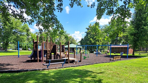 A playground with various play structures, including a slide, swings, and climbing frames, surrounded by grass and trees. - River Road Getaway (Muscoda)