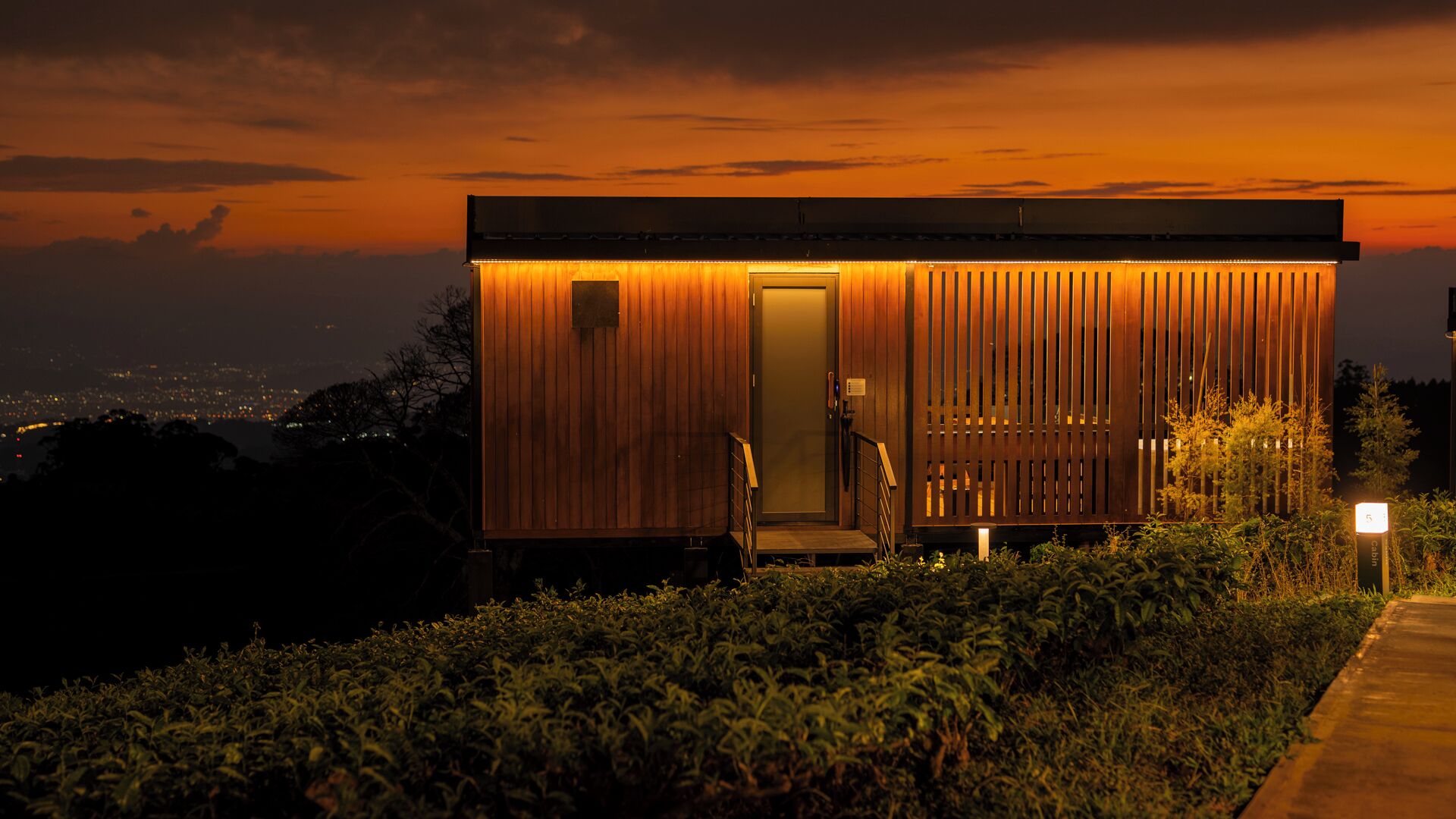 A modern, single-story building with a wooden facade and a glass door, surrounded by greenery.