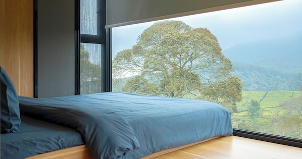 Ein Schlafzimmer mit großem Fenster, einem Bett mit blauer Bettwäsche und Blick auf Baum und Landschaft.