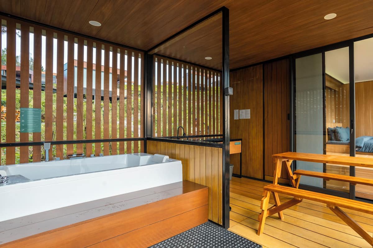 Un baño moderno con una bañera grande, piso de madera y una puerta de vidrio corredera que da a un dormitorio con cama y vistas al exterior.