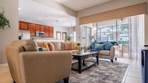 A spacious living room with a beige sofa, a glass coffee table, and a view of the kitchen and outdoor patio.