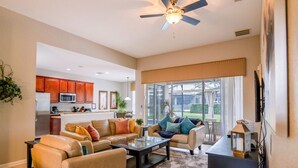 A spacious living room with a large sofa, a coffee table, and a television. There is a kitchen area with wooden cabinets and a ceiling fan.