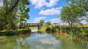Ein friedlicher Flussuferweg mit einer Holzbrücke, umgeben von üppigem Grün und einem Pavillon.