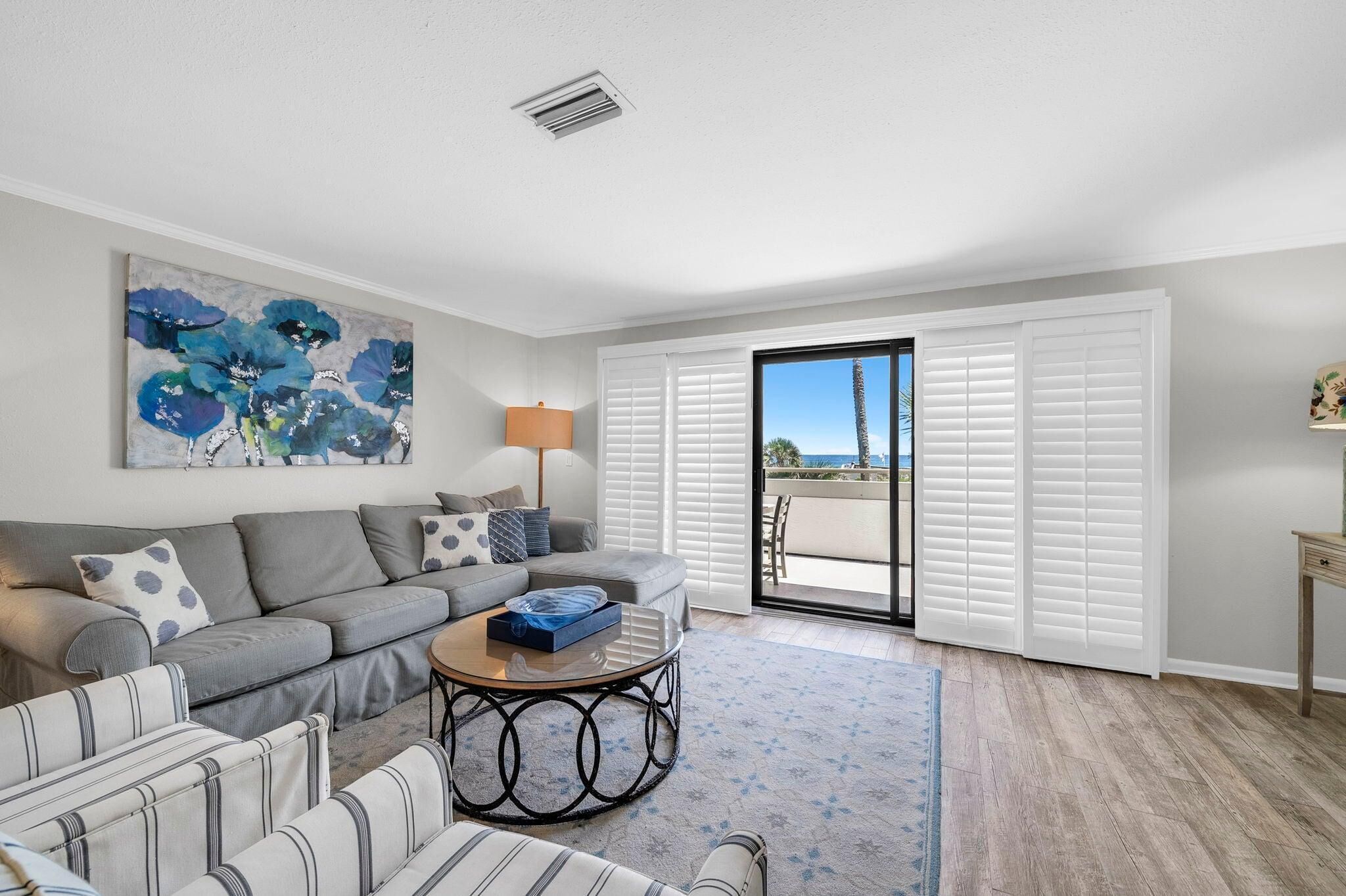 A living room with a sofa, armchairs, a coffee table, a painting, a lamp, and a sliding glass door leading to a balcony.